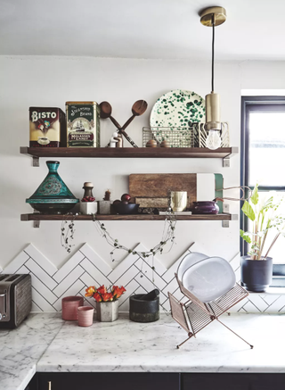 A white kitchen with marble worktops with a copper plate stand and decorative pots, a white herringbone splashback, and wooden shelving above it with decorative tins, plates, and chopping boards on it