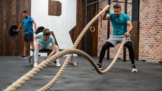 Three people in gym, man performing battle rope waves, woman performing concentration curl and man holding barbell