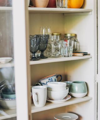An image of cream shelving unit with cups, plates, glasses, and other crockery mismatched on shelves