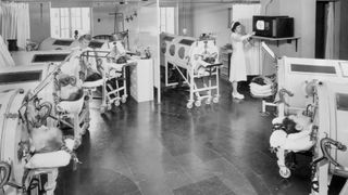 Black and white image of a group of eight patients who are in iron lungs that are arranged in an almost semi-circle manner around a television in a hospital. There are two nurses in the image, one is attending a patient towards the back of the image and another is standing by the television.