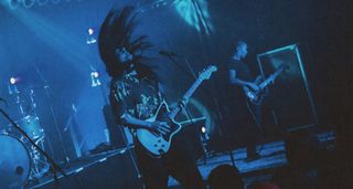 Neige of Alcest headbangs whille playing a hard-tail Jazzmaster onstage, in an image that is rendered in blue and black from the stagelights.