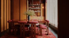 A dining room with chunky wood and red fabric chairs with a T-shaped back, a wooden table, a rug in pink and red tones, wrap-around wooden walls, see-through beige curtains, and a central flower vase.