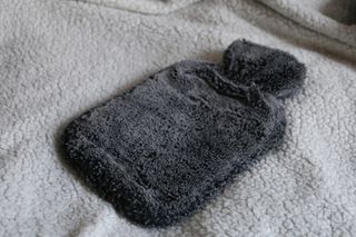 A dark grey, fluffy hot water bottle on top of a soft light grey blanket.