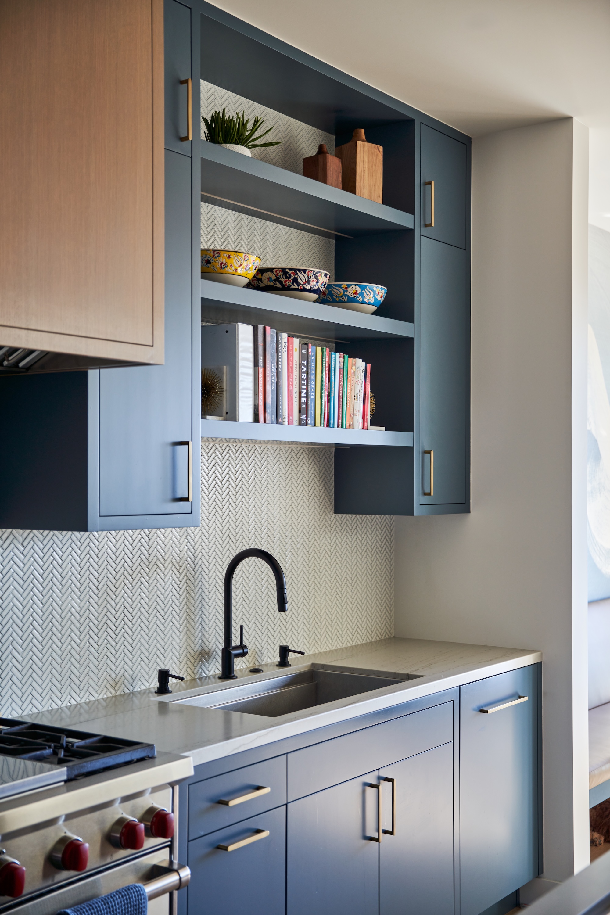 A deep blue kitchen sink area with a bllack tap and built in shelving above sink for decorative bowls and books