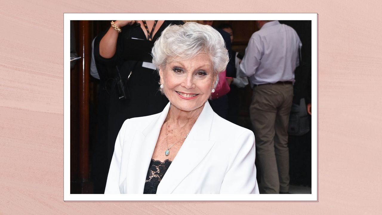 Portrait of Angela Rippon smiling wearing a white blazer with grey short hair, smokey eye and a coral lip, on a pink watercolour background