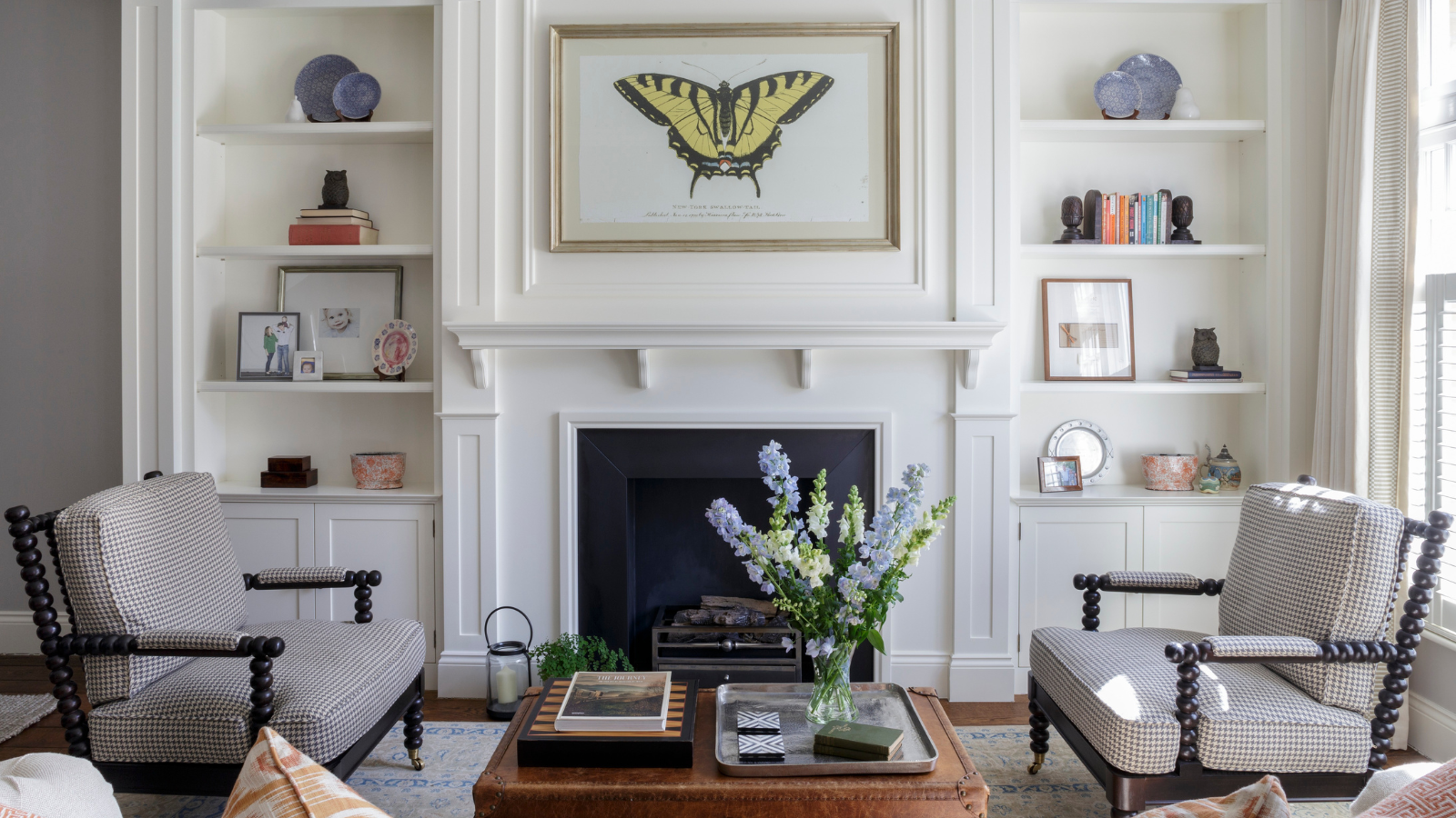 a symmetrical living room with a picture of a butterfly over the mantel, two accent chairs, a bouquet of flowers on the coffee table, and small decor in the bookshelves against the wall