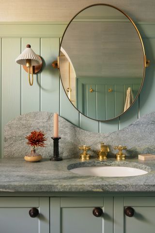 Close-up of bathroom vanity with stone countertop and backsplash, round mirror and traditional wall lights