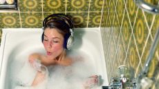 Young woman wearing headphones in bath blowing foam from hand, to illustrate self-care day ideas