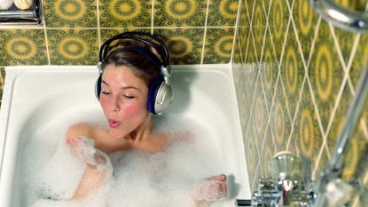 Young woman wearing headphones in bath blowing foam from hand, to illustrate self-care day ideas