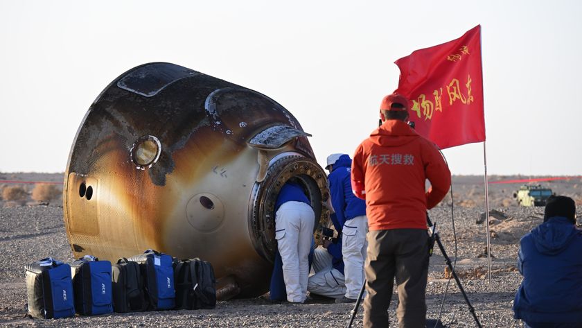 people in blue jackets lean into the opening of a charred cone-shaped spacecraft lying on its side on the rocky floor of a desert