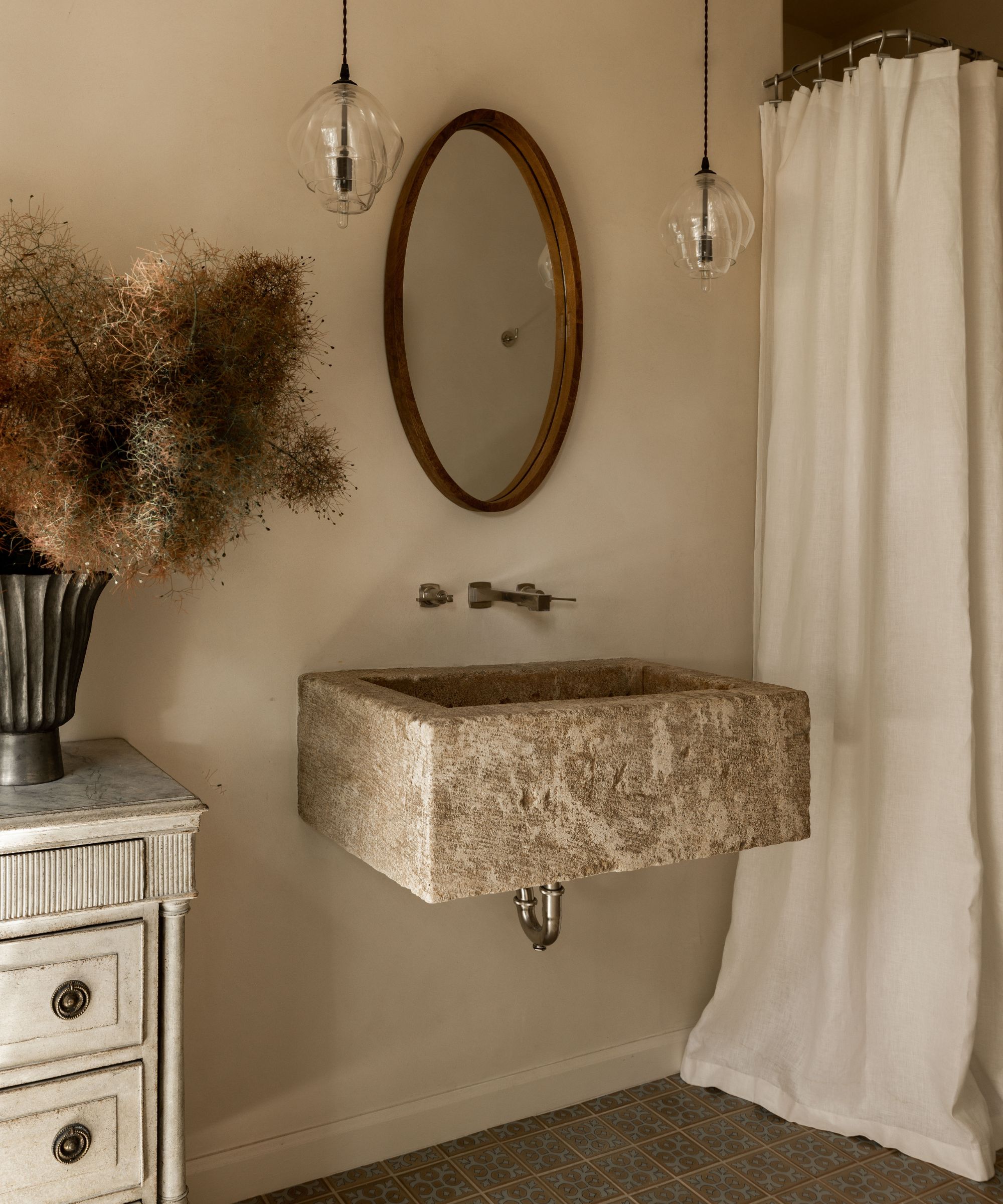 warm neutral small bathroom with a white linen shower curtain, stone floating sink, wooden mirror, and an antique dressers with a vase with branches