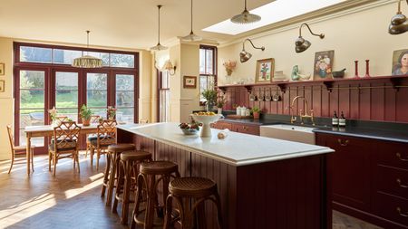 A large kitchen with the cabinets and island painted oxblood red, a paneled backsplash, black countertops on the perimeter cabinets, and a white marble counter on the island.