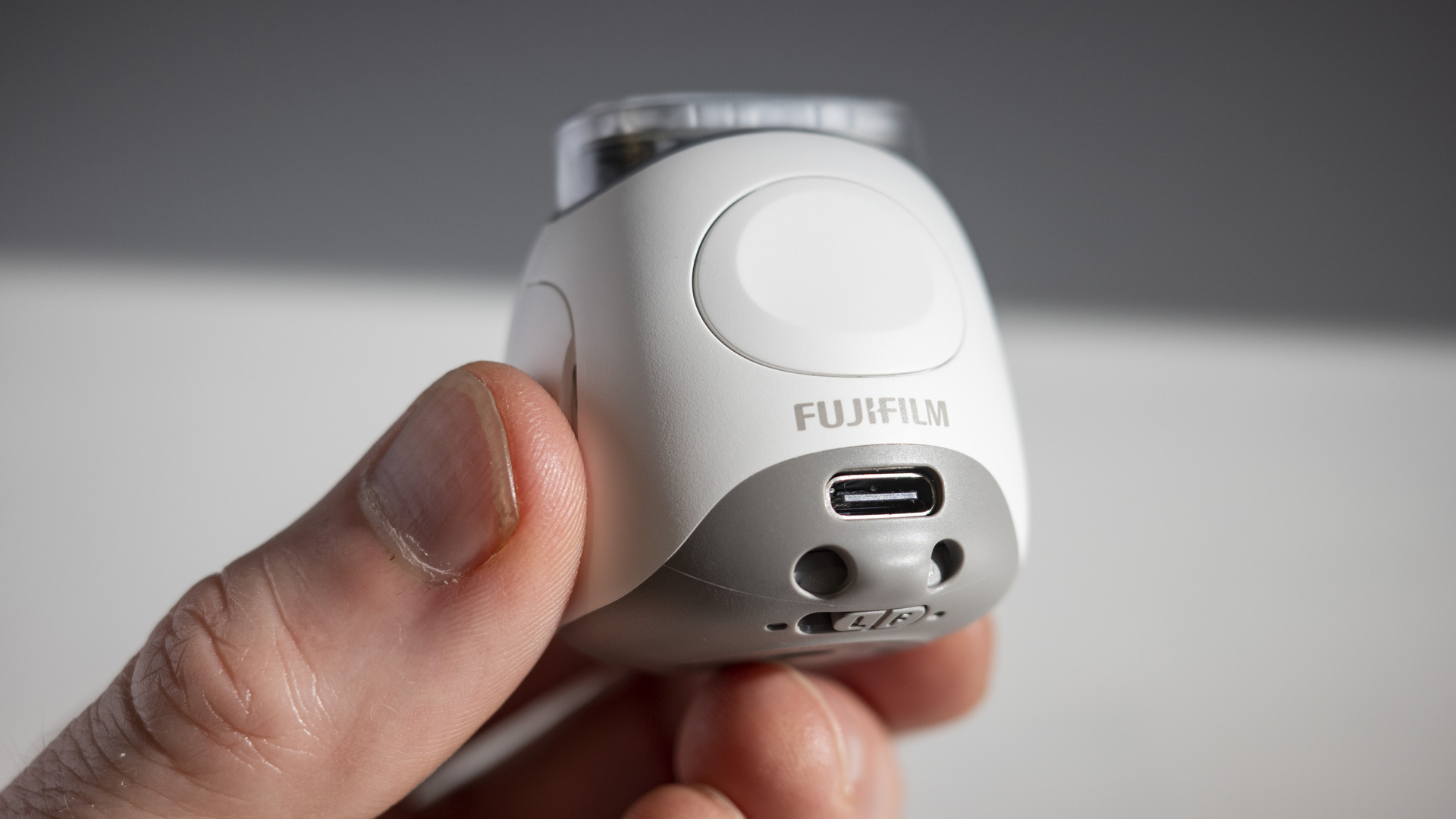 The rear side of a white Fujifilm Instax Pal, in the hand, with a white table in the background and harsh shadows