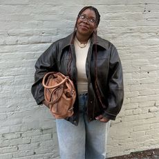 Chichi wears brown bomber, white tank, and denim jeans while holding a light brown leather tote bag