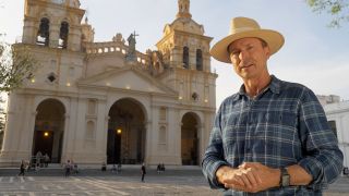 Phil Keoghan announces a challenge on location during The Amazing Race Season 36 in cool hat.