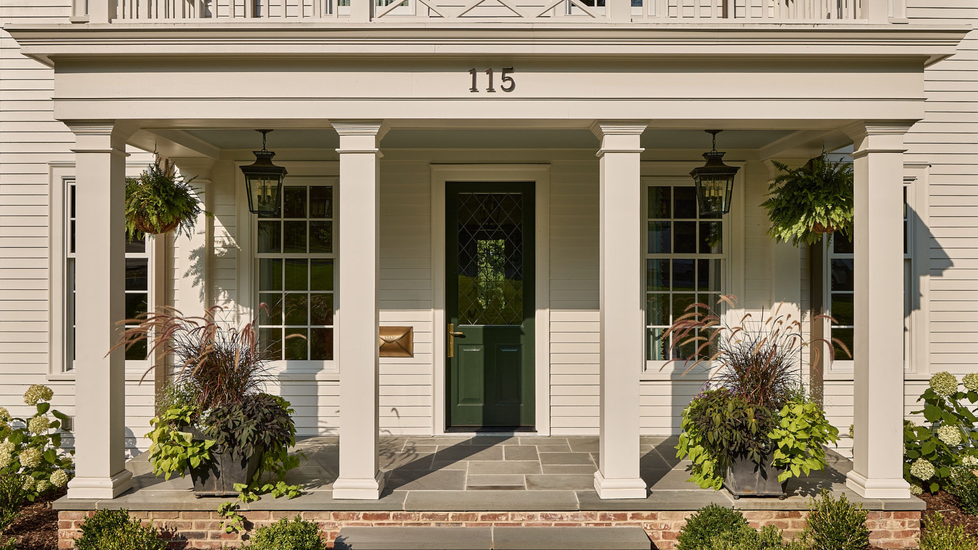 A large home exterior with warm white timber-clad walls, a front porch with four pillars, traditional-style windows and a dark green front door.