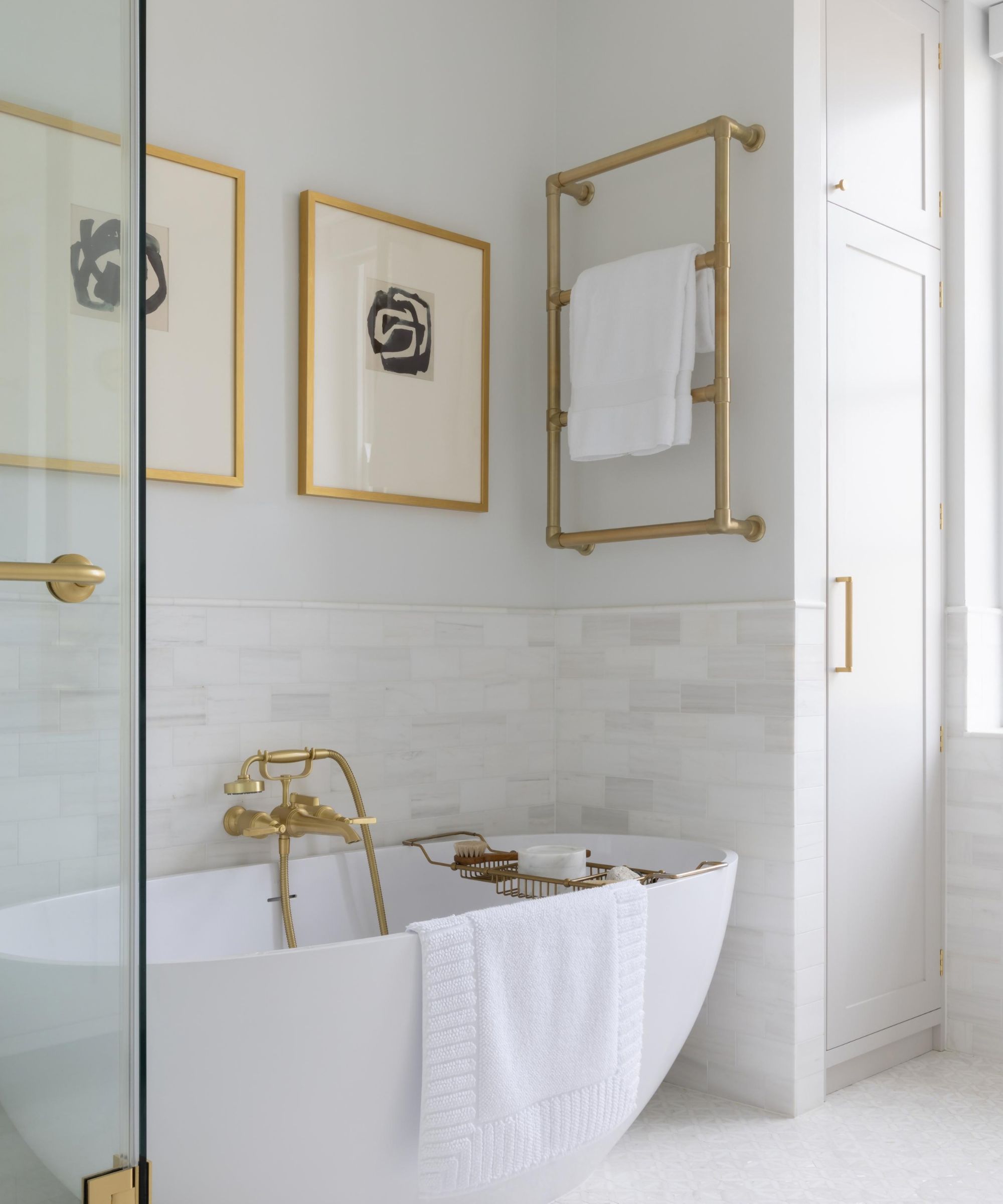 A bright, elegant bathroom with a white freestanding bathtub, brushed brass wall-mounted taps and a brass bath rack. A rectangular brass heated towel rail hangs above. White subway tiles, framed abstract artwork, built-in cabinetry and a glass shower enclosure complete the space.