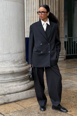 How to wear a suit is shown in a photo of a woman standing outside wearing oversized black eyeglasses with a black blazer styled with a silver earrings, a white button-down shirt, a white tie, black trousers, black loafers, and a black handbag