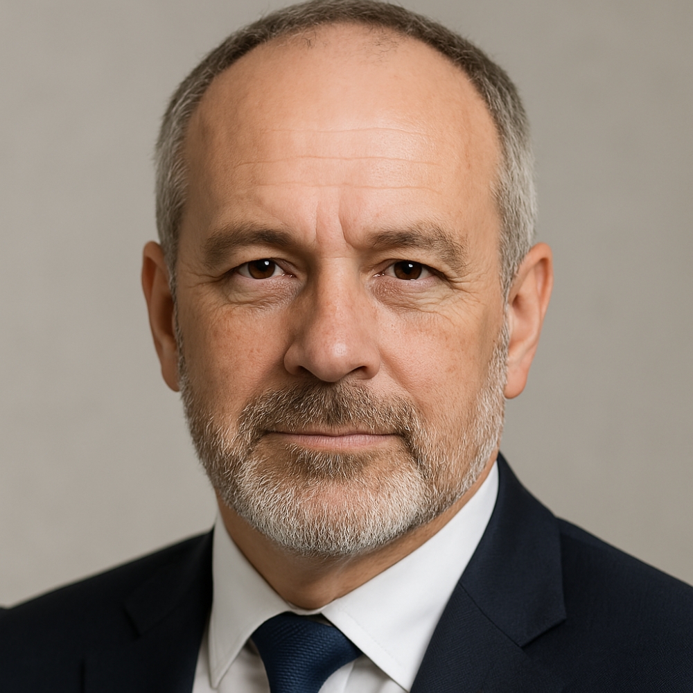Serious-looking older man with grey beard and hair, in a dark suit and blue tie, close-up headshot.