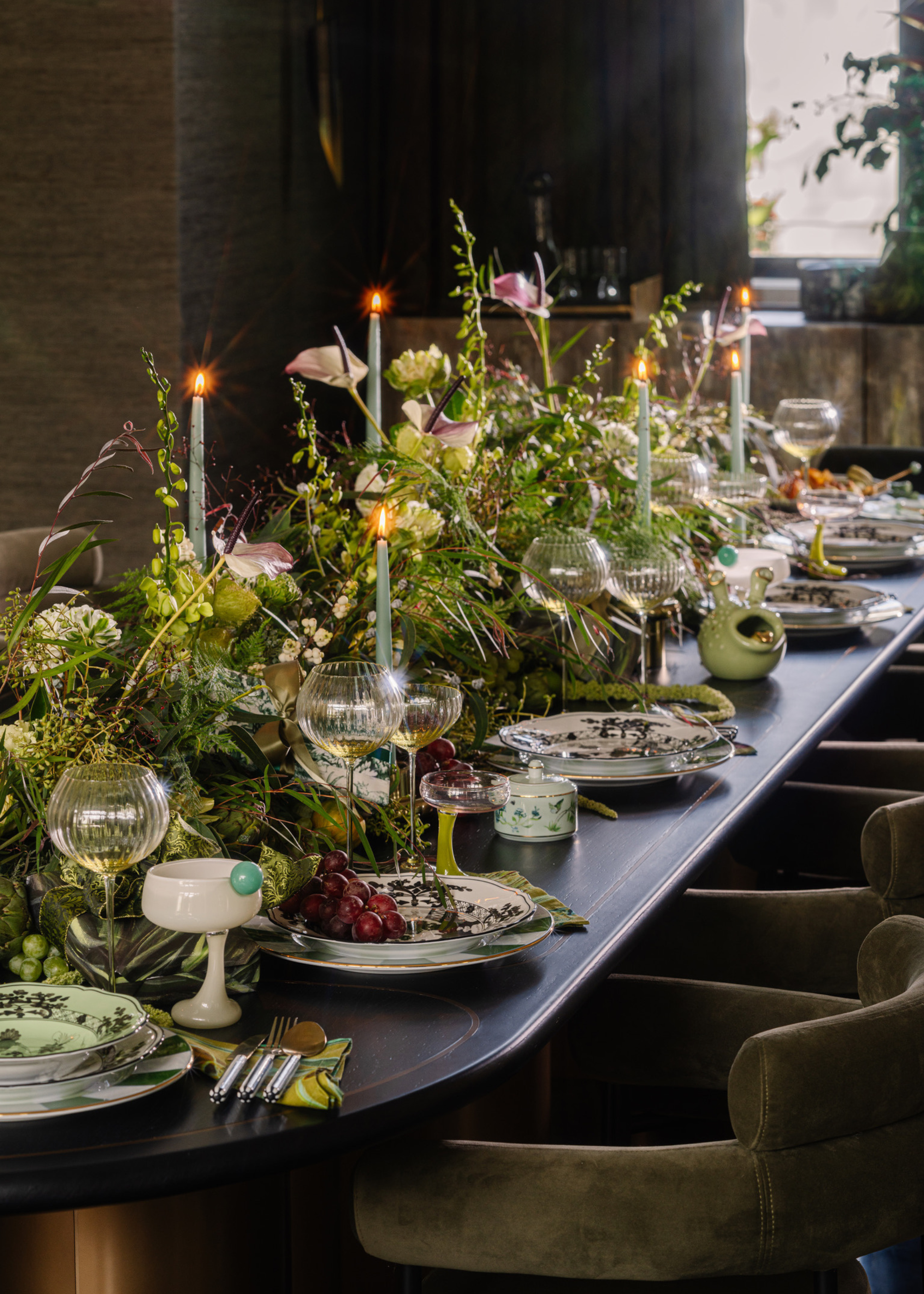 A Christmas table with a vibrant and layered centerpiece that runs the length of the table, with foliage and lit taper candles