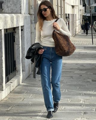 French fashion influencer Anne-Laure Mais walking on a Paris sidewalk with black sunglasses, a sheer beige longe-sleeve t-shirt, leopard-print shoulder bag, leather jacket draped on her arm, cuffed jeans, and black boots.