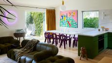 A white living room with mustard window treatments, a colorful painting, a white dining table with purple dining chairs, a dark green sofa set with a striped throw, a purple backlit sconce, a pendant light, a green kitchen with white counters, and a wood stool