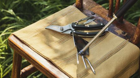 Shears and a hand rake on a taupe cloth draped across a wooden chair in a garden