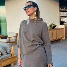 Sofia richie wears black sunglasses, hanging earrings, a gray sweater, and a gray knit skirt.