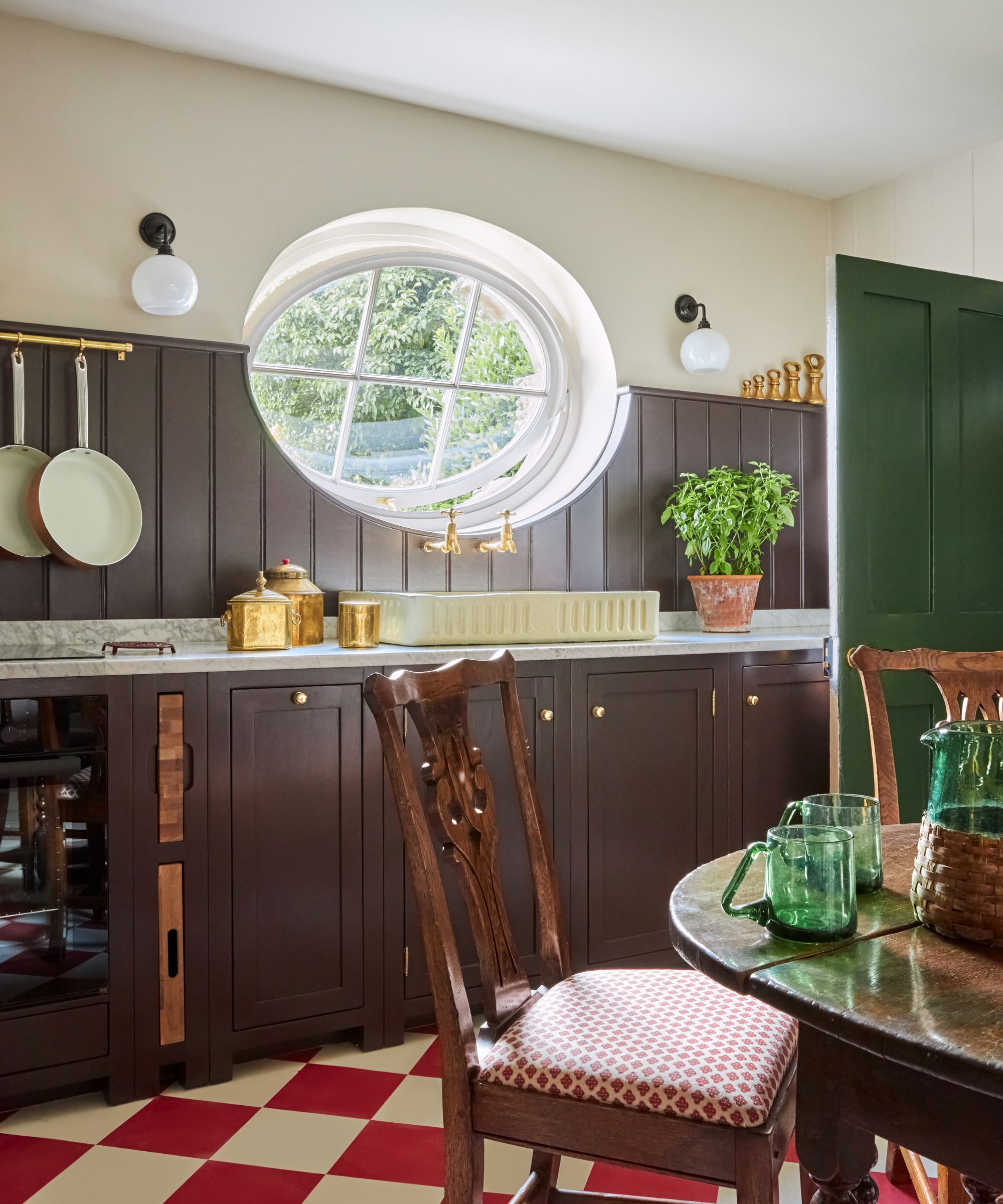 Rustic kitchen featuring dark brown wood paneling, white marble countertops, a cream-colored sink under a large oval window, and wooden dining chairs in the foreground