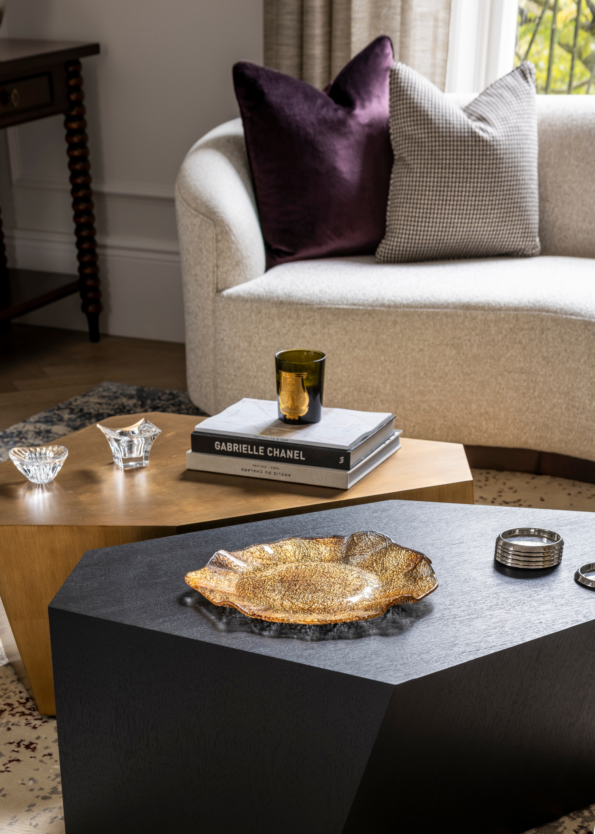 Black and natural wood coffee tables in irregular shapes are styled next to one another in a living room with a beige sofa with a decorative tray, objects, candle and books