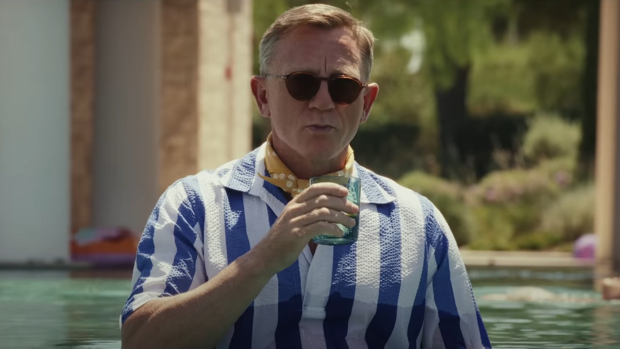 Daniel Craig como Benoit Blanc de pie en una piscina con una bebida y con gafas de sol en Glass Onion: A Knives Out Mystery