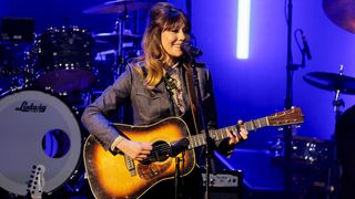 Molly Tuttle playing acoustic guitar on stage, with a drum kit in the background