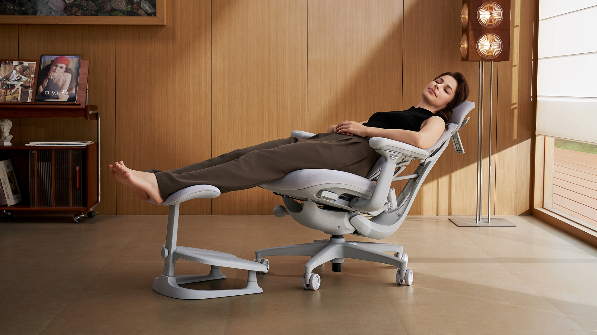 A woman reclines fully in a light grey ergonomic office chair with a footrest in a warm, wood-panelled room, with sunlight streaming in from the right.