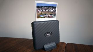 En svart Fujifilm Instax Link Wide står på ett mörkt träbord och har precis skrivit ut ett foto.