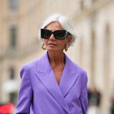 Grece Ghanem wears black sunglasses from Saint Laurent, a beige leather and gold braided earrings, a purple oversized blazer jacket, matching purple large suit pants, a dark brown shiny braided leather handbag, outside the COS show, on April 26, 2023 in Paris, France