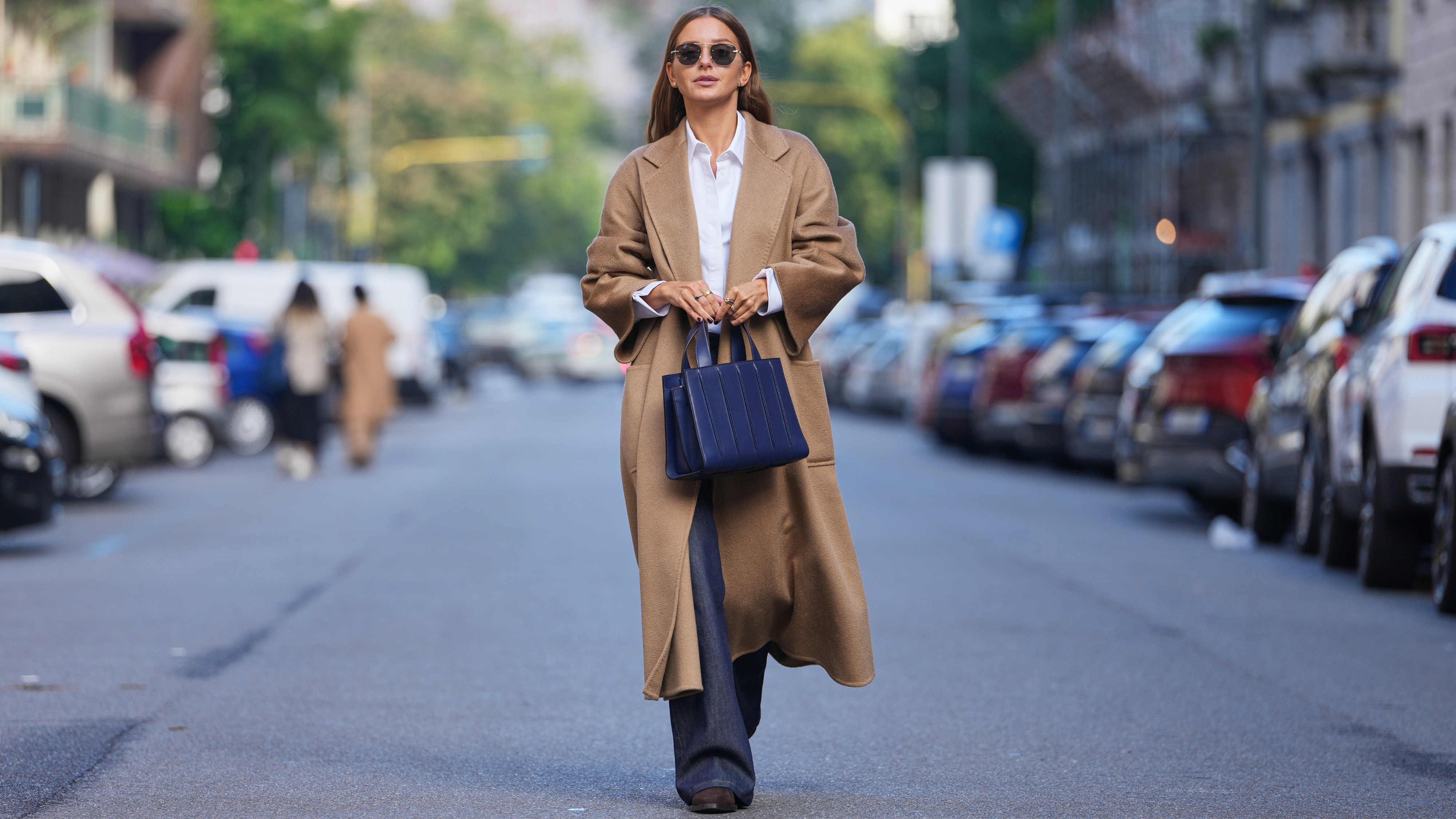 Diane Batoukina wears long straight brown hair with a center part, round black sunglasses, gold rings, a navy blue leather tote bag, a camel wool long coat, a white cotton button-up shirt, dark indigo denim wide-leg jeans, black leather boots shoes with a low block heel, outside Max Mara, during Milan Fashion Week