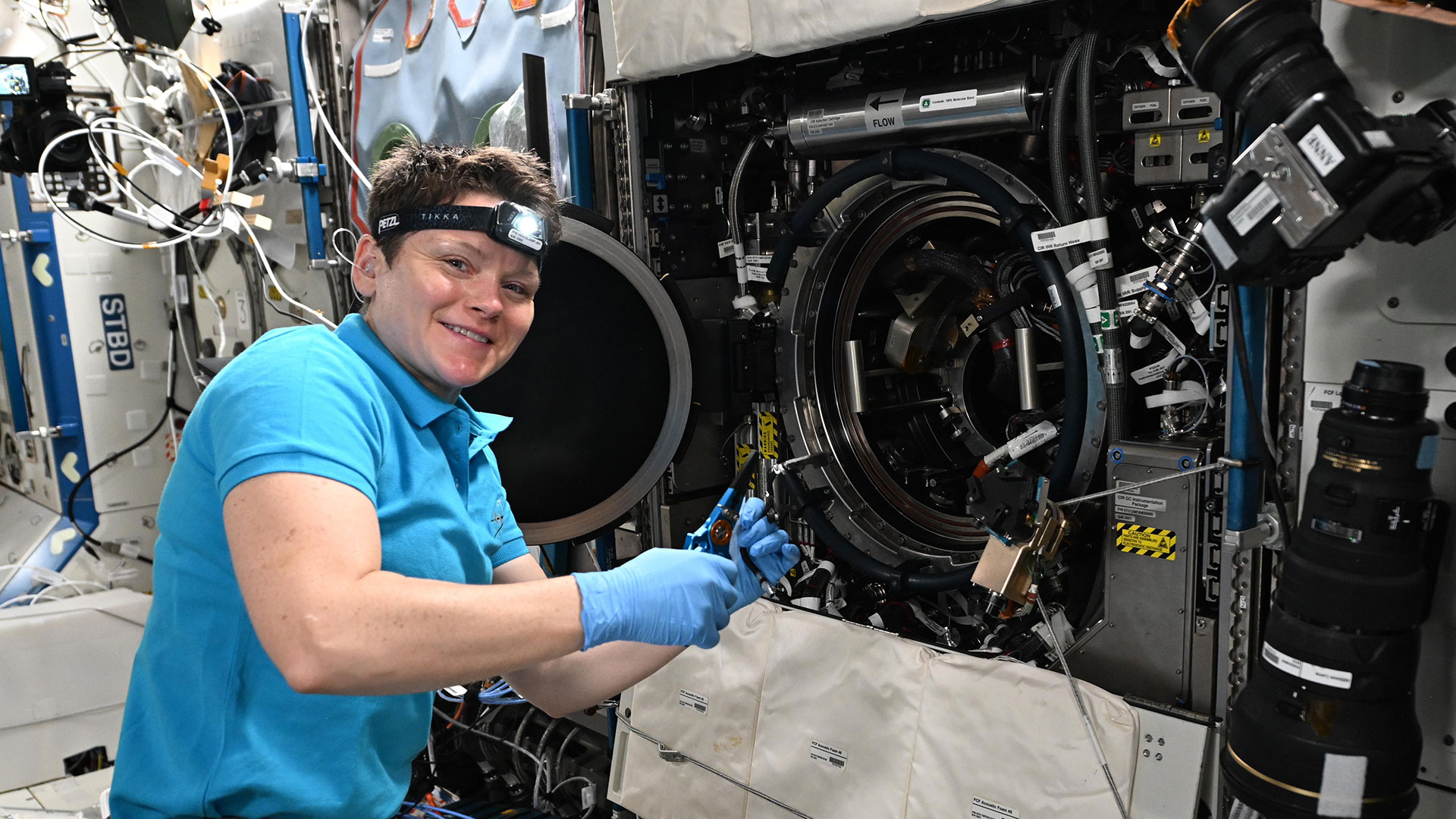 a woman wearing a blue polo shirt and headlamp works next to a science experiment aboard a space station