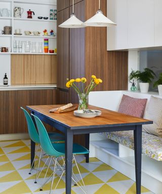 Kitchen diner with yellow and white, geometric vinyl flooring, dark wood cupboards and dining table, bench seating with cushions, blue dining chairs, low hanging pendants