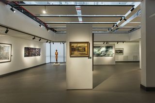 Interior view of the Javett Art Centre gallery featuring white walls, grey flooring, black bar mounted spotlights, art on the walls and a sculpture of a woman on a low, white plinth