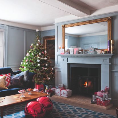 A baby blue-painted living room with a fireplace decorated for Christmas with the Christmas tree tucked in the corner and presents laid out underneath it