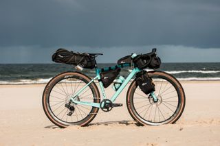 The Jack Wolfskin bikepacking luggage is all loaded on to a blue bike that is on the sand with the sea in the background