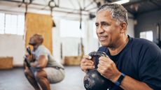 Two men in a gym squat down and hold a kettlebell to their chest.