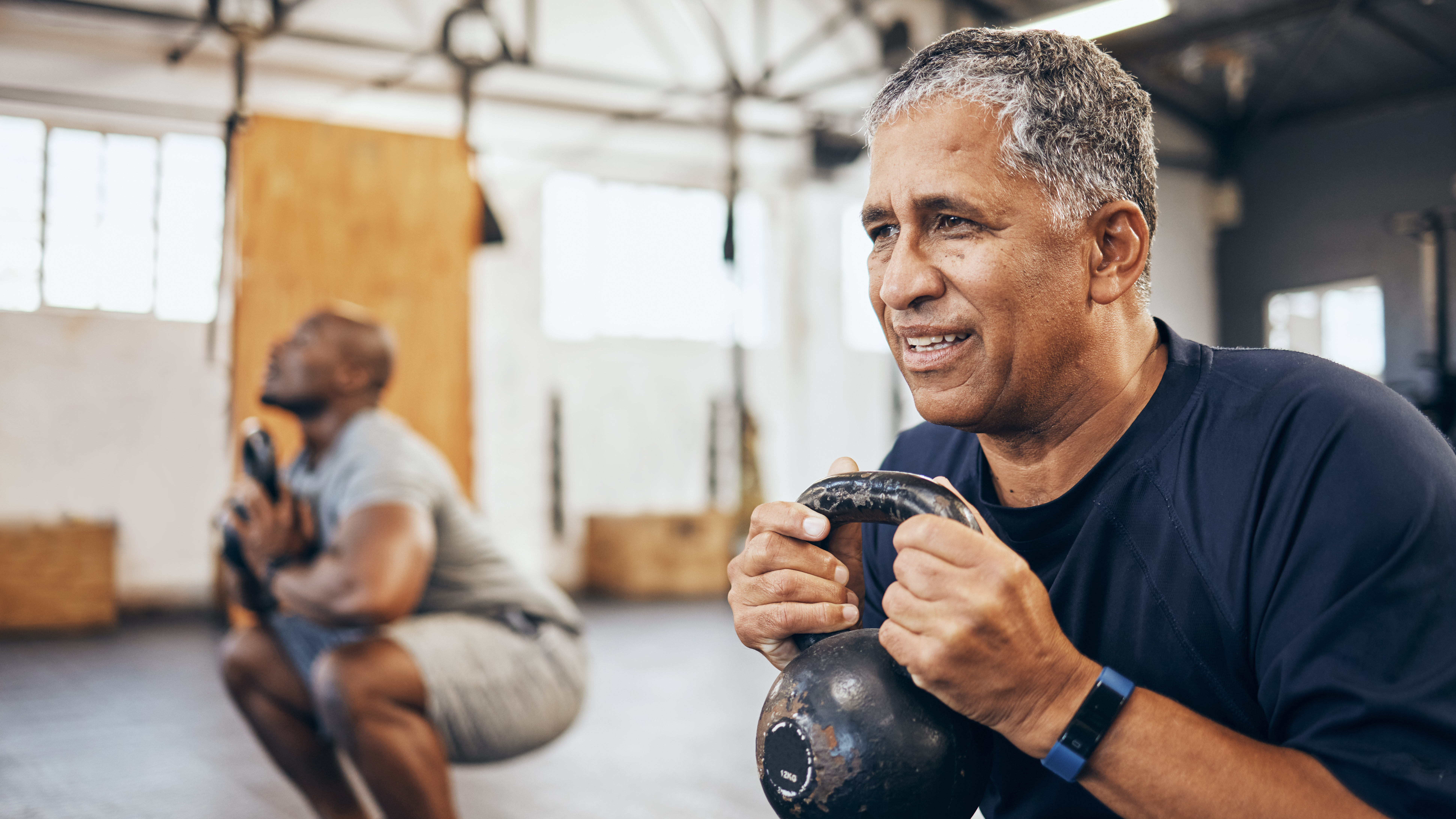 Two men in a gym squat down and hold a kettlebell to their chest.