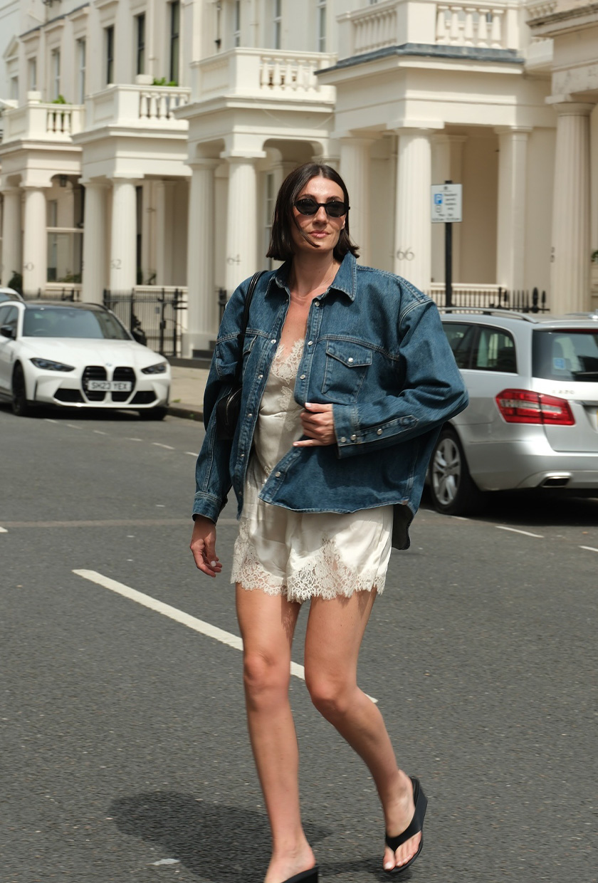 The sandal trend wedge flip-flops are shown in a photo of a woman crossing the street, wearing a denim shirt on top of a white satin camisole and pajama shorts with a black shoulder bag, black sunglasses, and wedge flip-flops