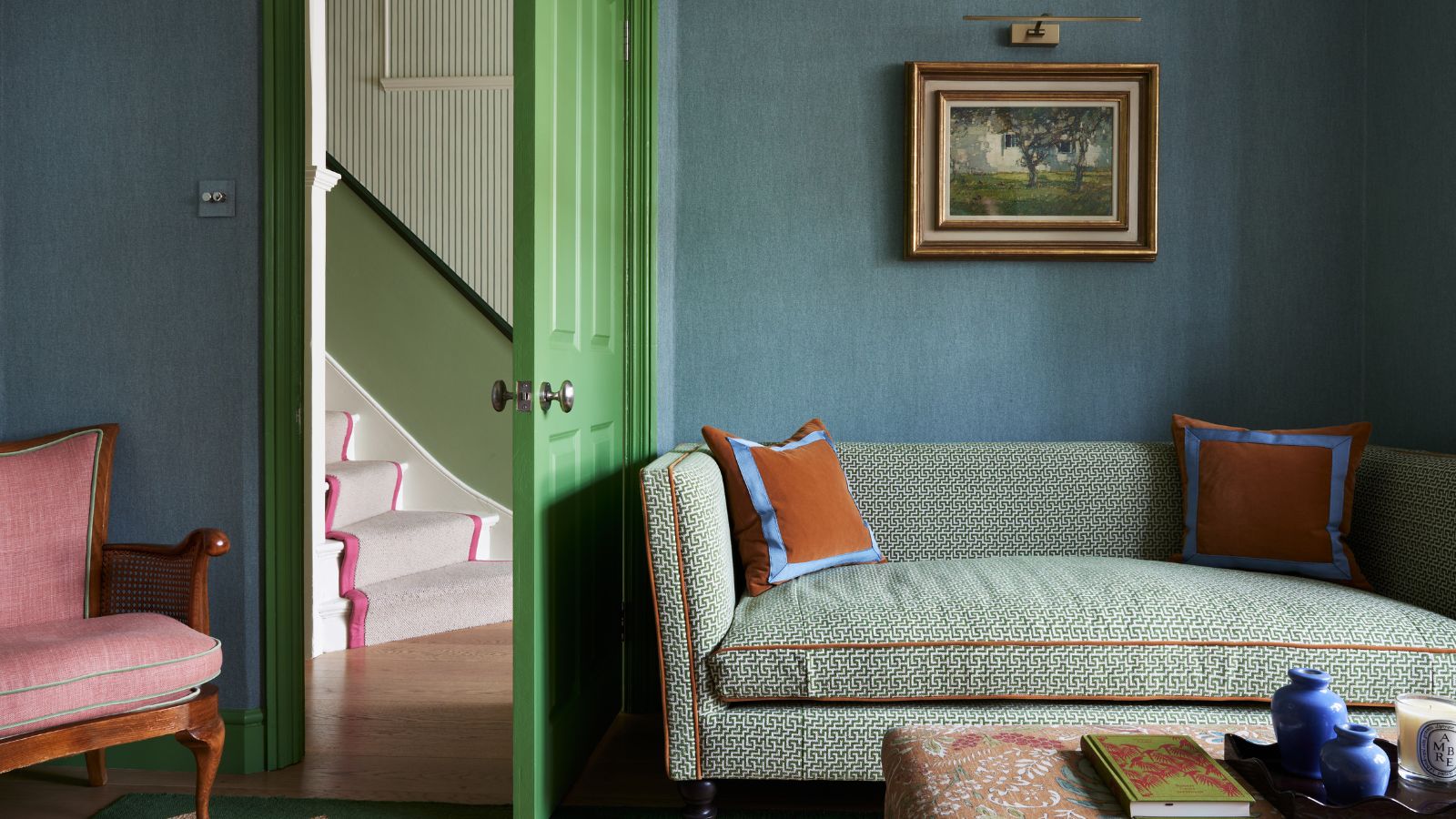 Living room with blue grasscloth walls, a green painted door, a green sofa with orange piping, orange velvet cushions, a coffee table with books and vases, and a pink armchair in the corner.