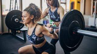 Woman performs back squat with barbell in gym. Another woman stands behind her, hands hovering under the bar ready to help