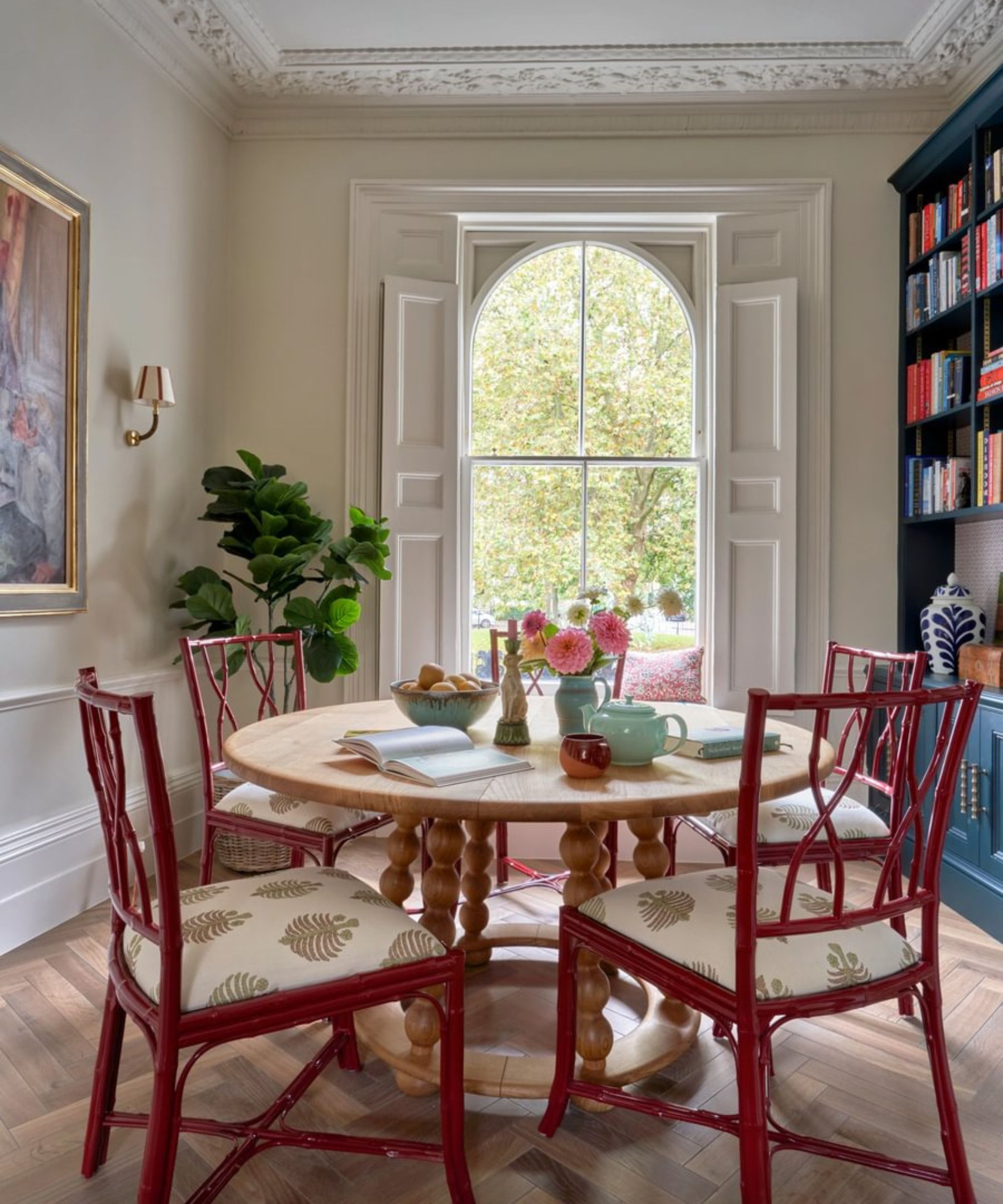 neutral victorian dining room with arched window and shutters, a round wooden table with bobbin legs, four bamboo style lacquer chairs, and a blue bookcase