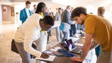 A photo of a student stood signing a registration form at a campus event, as a tech organizer looks on. On the table the form is on, there are lanyards and a laptop. Decorative: the student has dark skin and wearing a white jacket, while the tech organizer has medium-light skin and is wearing a mustard yellow polo shirt.