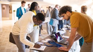 A photo representing campus recruitment of a student stood signing a registration form at a campus event, as a tech organizer looks on. On the table the form is on, there are lanyards and a laptop. Decorative: the student has dark skin and wearing a white jacket, while the tech organizer has medium-light skin and is wearing a mustard yellow polo shirt.