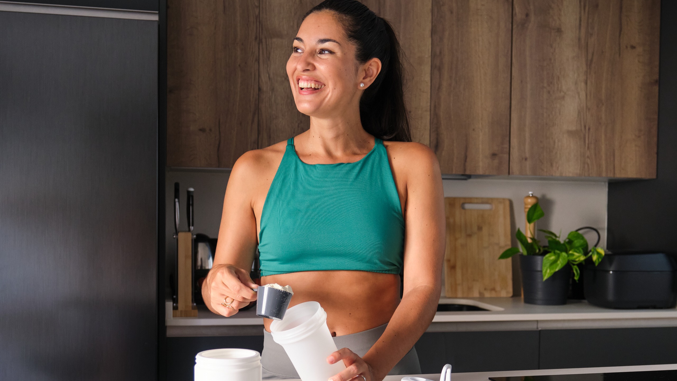 A woman is stood in her kitchen with a scoop of a powdered supplement in one hand and a cup in the other. 